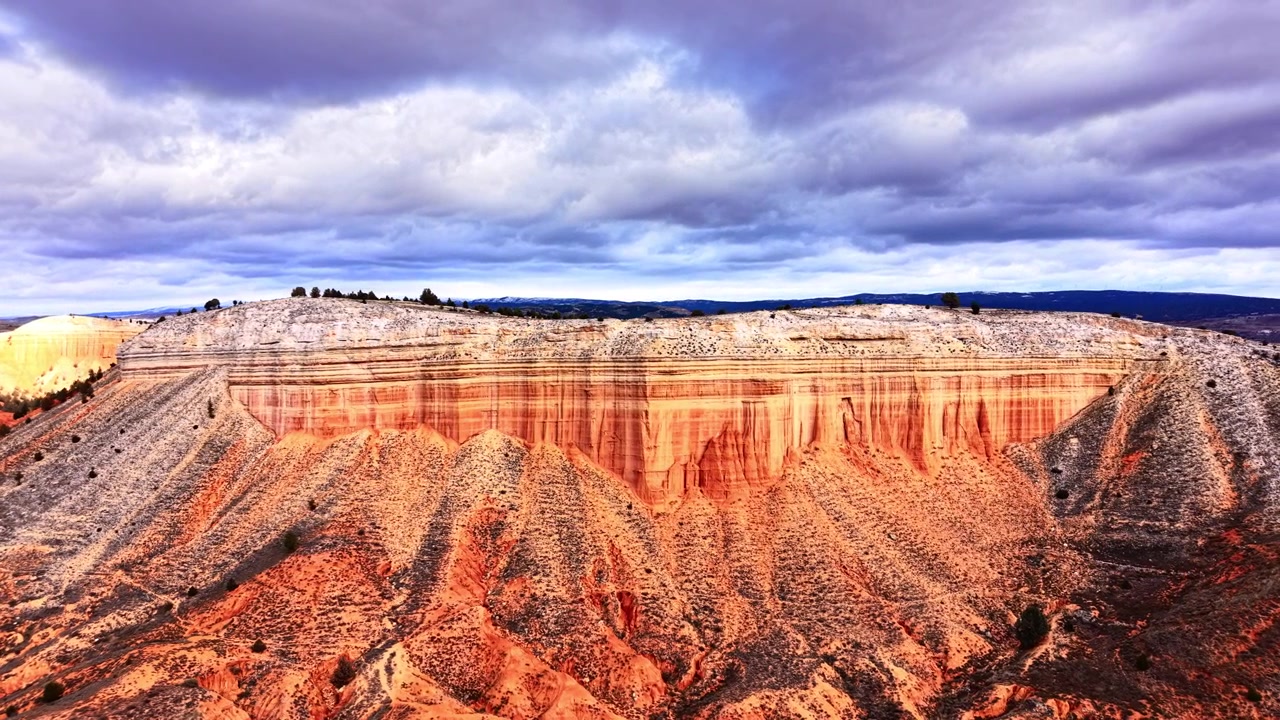 Ampia vista di una mesa di arenaria rossa erosa sotto un cielo nuvoloso drammatico, Spagna. Altopiano stratificato con cima piatta, strato roccioso arancione e grigio, calanchi scolpiti in primo piano, arbusto sparso