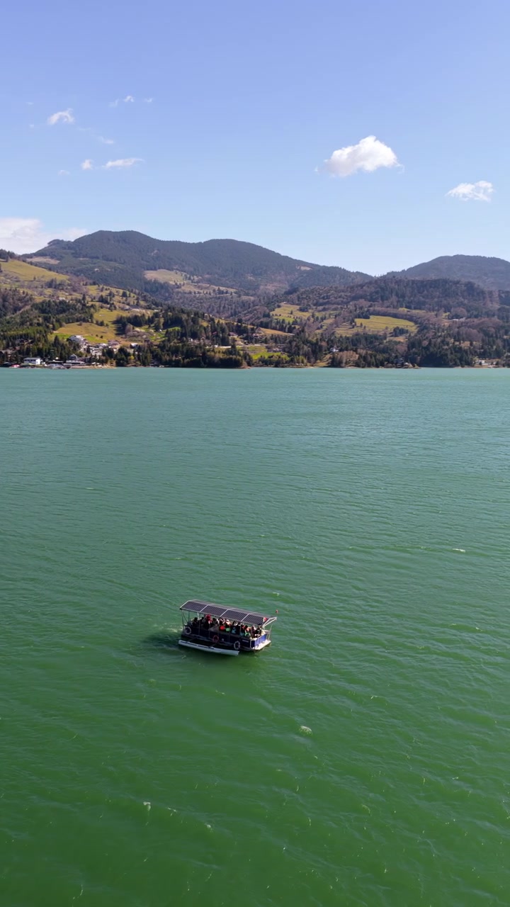 Vista aerea con drone di una piccola imbarcazione passeggeri che attraversa il lago Colibita con montagne sullo sfondo, in Romania. Verticale