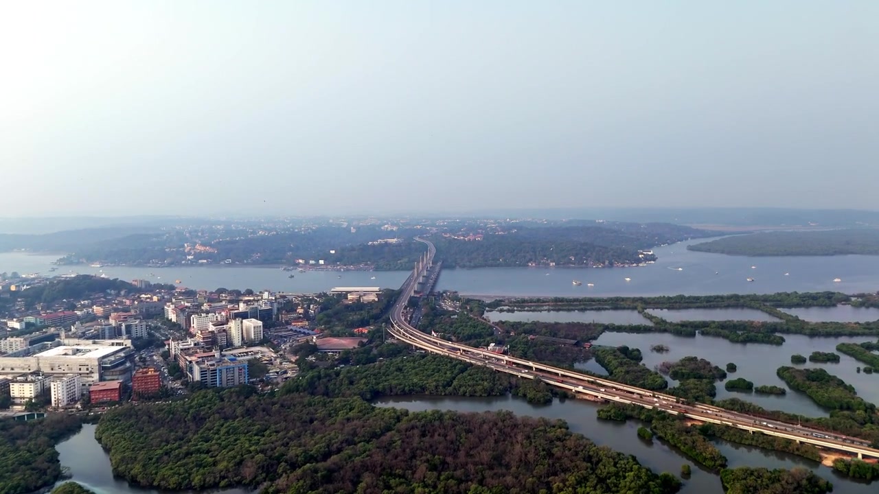 Ripresa aerea con drone che cattura il ponte Atal Setu che attraversa l'ampio fiume Mandovi, mostrando un flusso di traffico scorrevole, corsi d'acqua a più livelli e una leggera foschia atmosferica con formazioni terrestri distanti che scompaiono gradualmente.