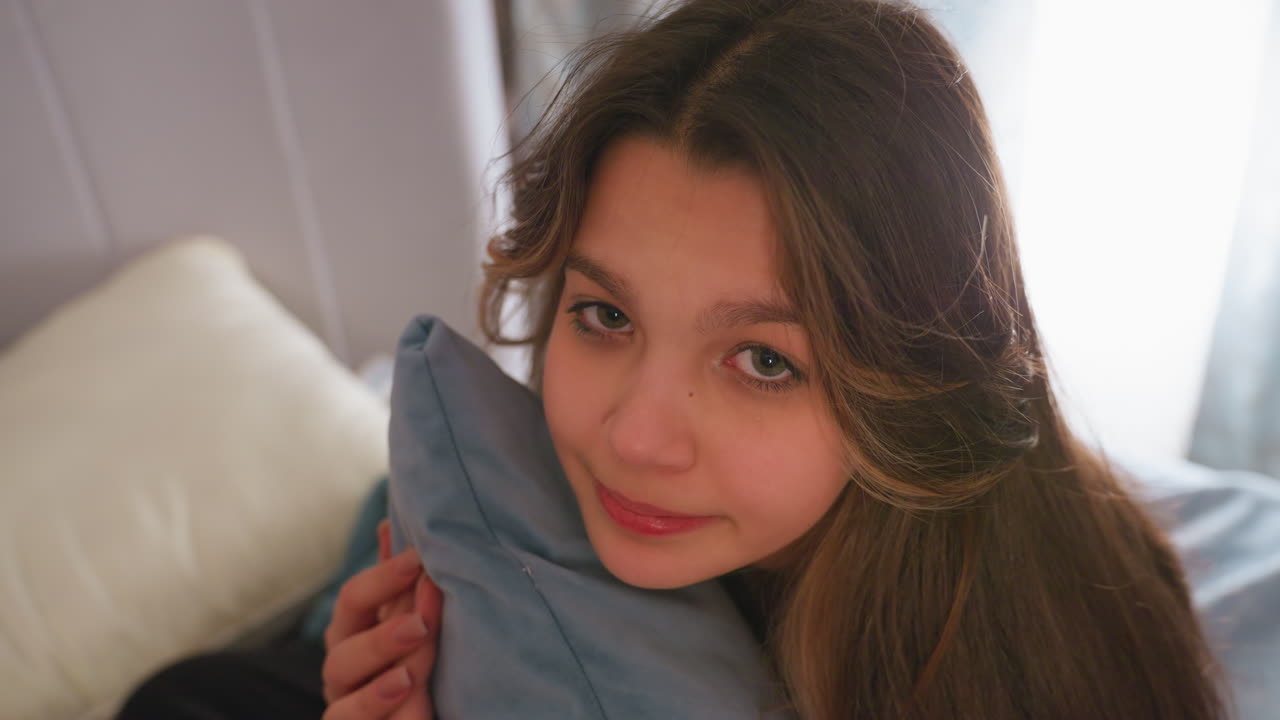 Young woman with long brown hair gently hugs soft blue pillow while gazing forward with an innocent expression, warm lighting and soft bedding