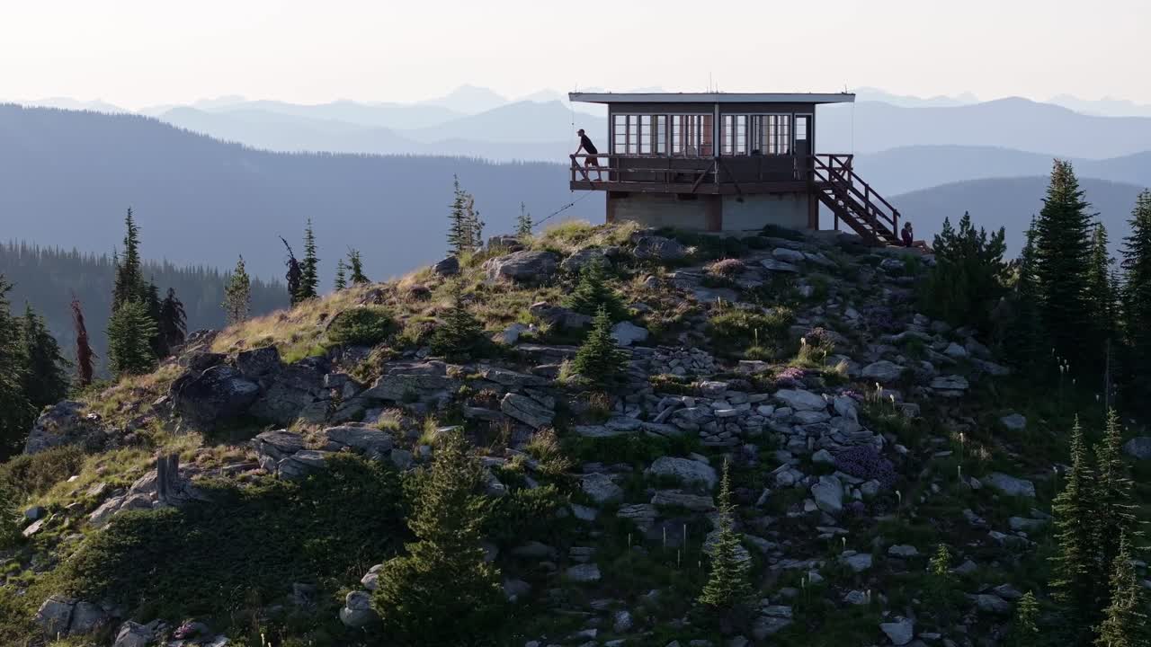 En mand går i slowmotion fra højre mod venstre ud på verandaen og læner sig op ad rækværket på et isoleret brandtårn på en bjergtop i det fjerntliggende nordlige Idaho foran et barsk landskab