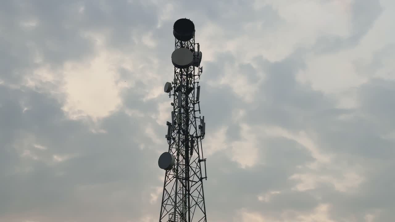 A cinematic tilt-up shot of a tall communication tower set against a soft morning sky with clouds drifting, highlighting technology blending with natural surroundings at sunrise