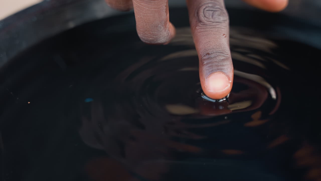 Dito che tocca una macchia scura in un secchio, primo piano di un test di finitura per falegnameria che mostra la punta del dito creare leggere increspature su un liquido lucido, riflesso e ombra sottili, ispezione tattile del campione di macchia.