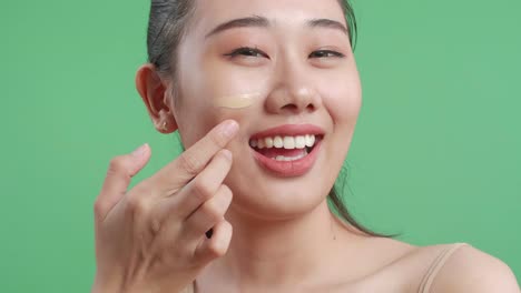 스튜디오에서 녹색 스크린 배경에 서서 얼굴 크림을 적용하고 미소 짓는 아시아 여성의 클로즈업