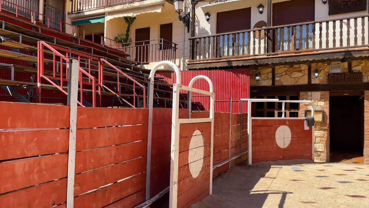 We see a mobile bullring installed in a town square with festive motifs, we see the burladero with a white circle, the structure of the bullring is made of wood and iron, there is dirt on the ground