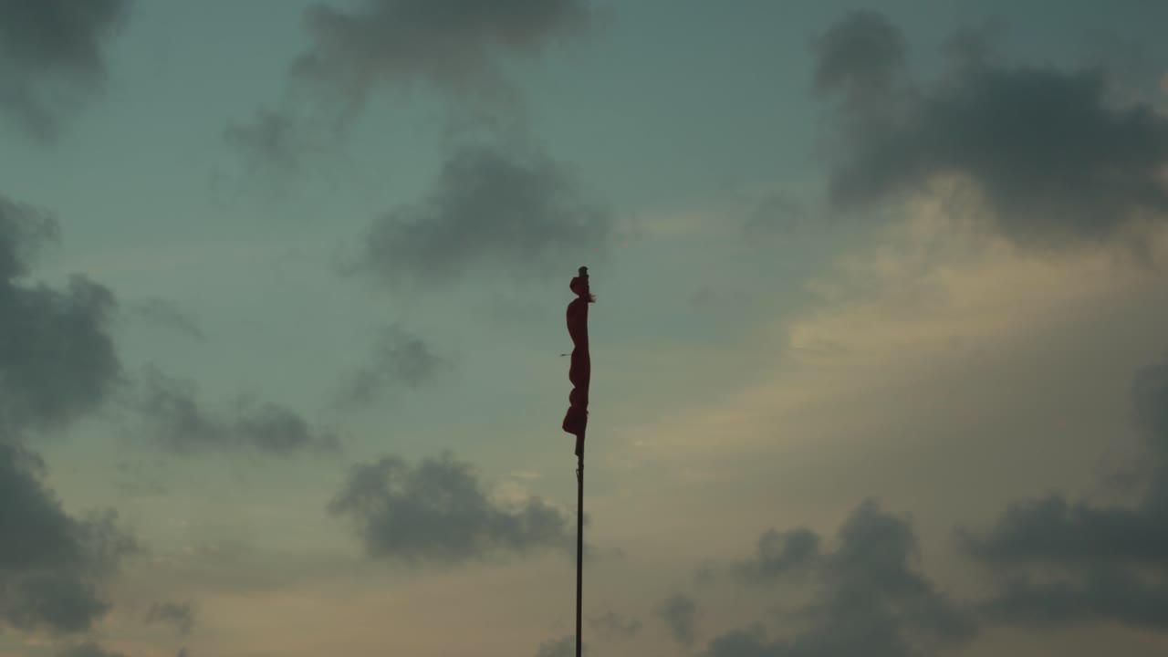 Rødt flag, der blafrer i vinden på en høj stang med skyer og dramatisk solnedgangshimmel, minimal scene, koncept for advarsel, symbolik og atmosfære.
