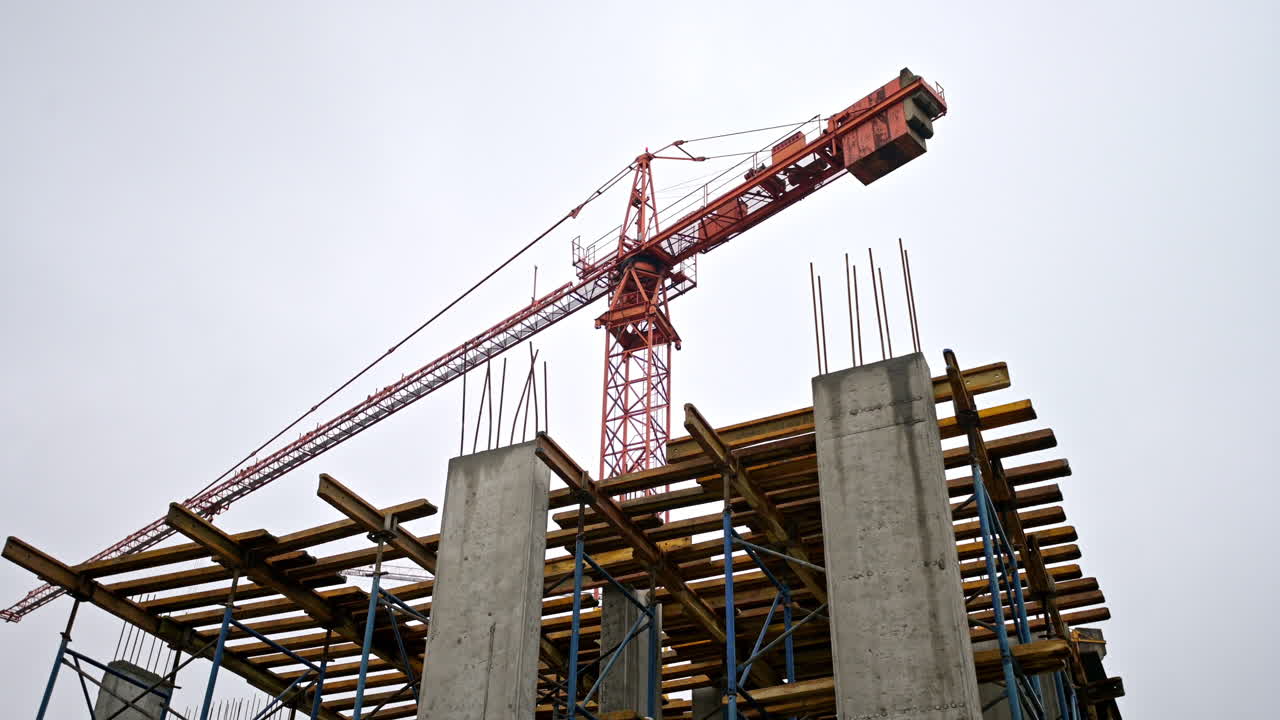 Low angle view of a red crane operating on a concrete structure. Professional building process with reinforced pillars and formwork under a cloudy sky. Modern urban engineering project