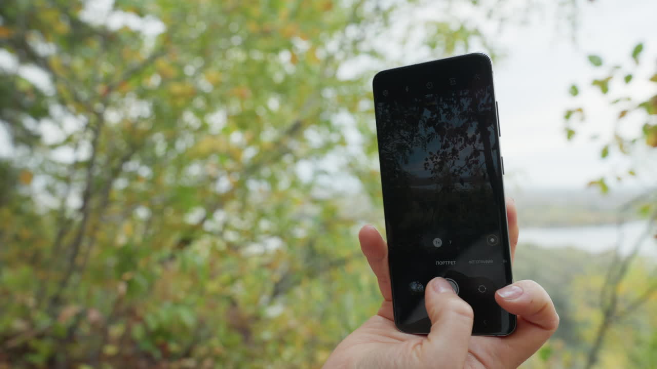 Registrazione con telefono portatile della riva di un fiume panoramico in stile vlogger: donna tiene il telefono in orizzontale, inquadra i rami mossi sull'acqua, tocca registra, il pollice stabilizza l'inquadratura, contenuto naturalistico intimo
