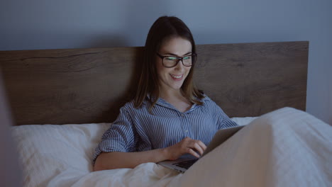 smiling caucasian young woman in eyeglasses typing on the keyboard of the laptop late at night in the bed