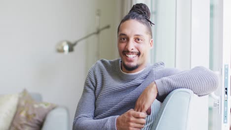 portrait of happy biracial man with dreadlocks in hair bun sitting on couch smiling in living room