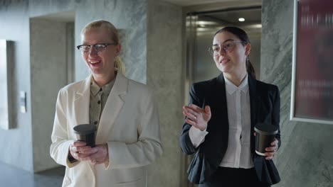 Confident-blonde-girl-in-a-white-business-suit-walks-with-her-brunette-female-colleague-during-the-start-of-their-working-day-and-coffee-break-in-a-modern-office