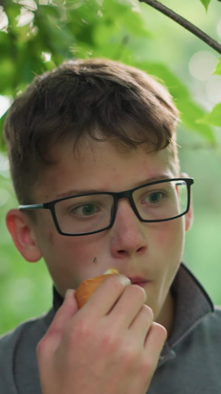 joven con una camiseta gris y gafas comiendo un bocadillo tranquilamente bajo un árbol, aparece profundamente en pensamiento, rodeado de hojas verdes exuberantes en un ambiente al aire libre tranquilo
