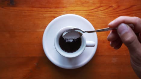 Toma-De-Una-Mano-Que-Está-Poniendo-Azúcar-En-Una-Taza-De-Café-Expreso-Caliente-Y-Se-Agita,-Sobre-Una-Mesa-De-Madera-Marrón