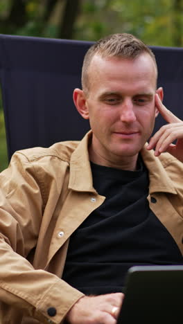 pensive sitting in garden chair looks thoughtfully at his laptop screen. freelancer working in nature has found some mistake and looks closer at computer. vertical video