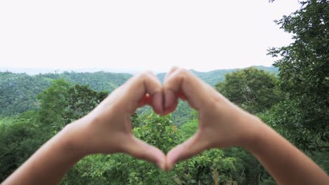 manos con los dedos unidos formando un corazón revelando las montañas y el bosque alejado mostrando tierras y la cabeza de una mujer