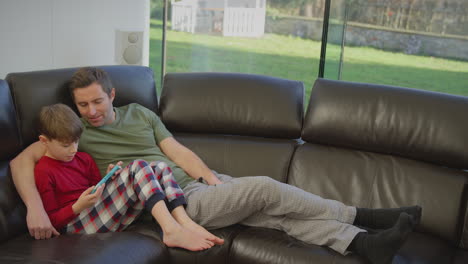 father with son wearing pyjamas lying on sofa at home playing computer game on handheld device - shot in slow motion