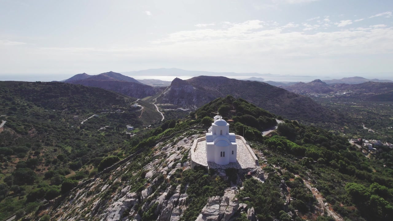 Sorvolo aereo con drone 4K della cappella del Profeta Elia sulla cima di Naxos, Grecia. Cupola cicladica imbiancata a calce contro drammatici dirupi rocciosi e pendii montuosi