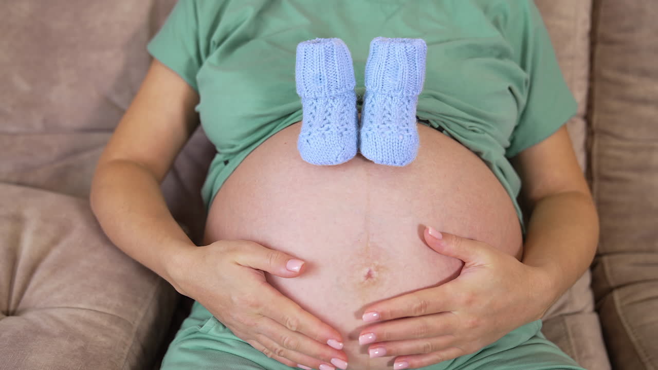 Future mum caress her big belly. Blue knitted socks on a bare stomach pf a pregnant woman. Women's health and people concept. Maternity prenatal care.
