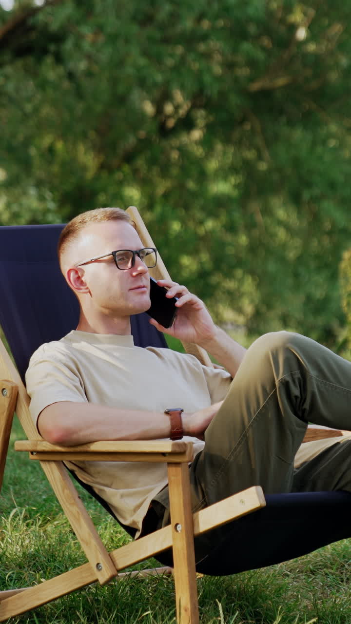 Serious Caucasian male sits in a folding chair having phone conversation. Man combining work and rest in nature. Vertical video