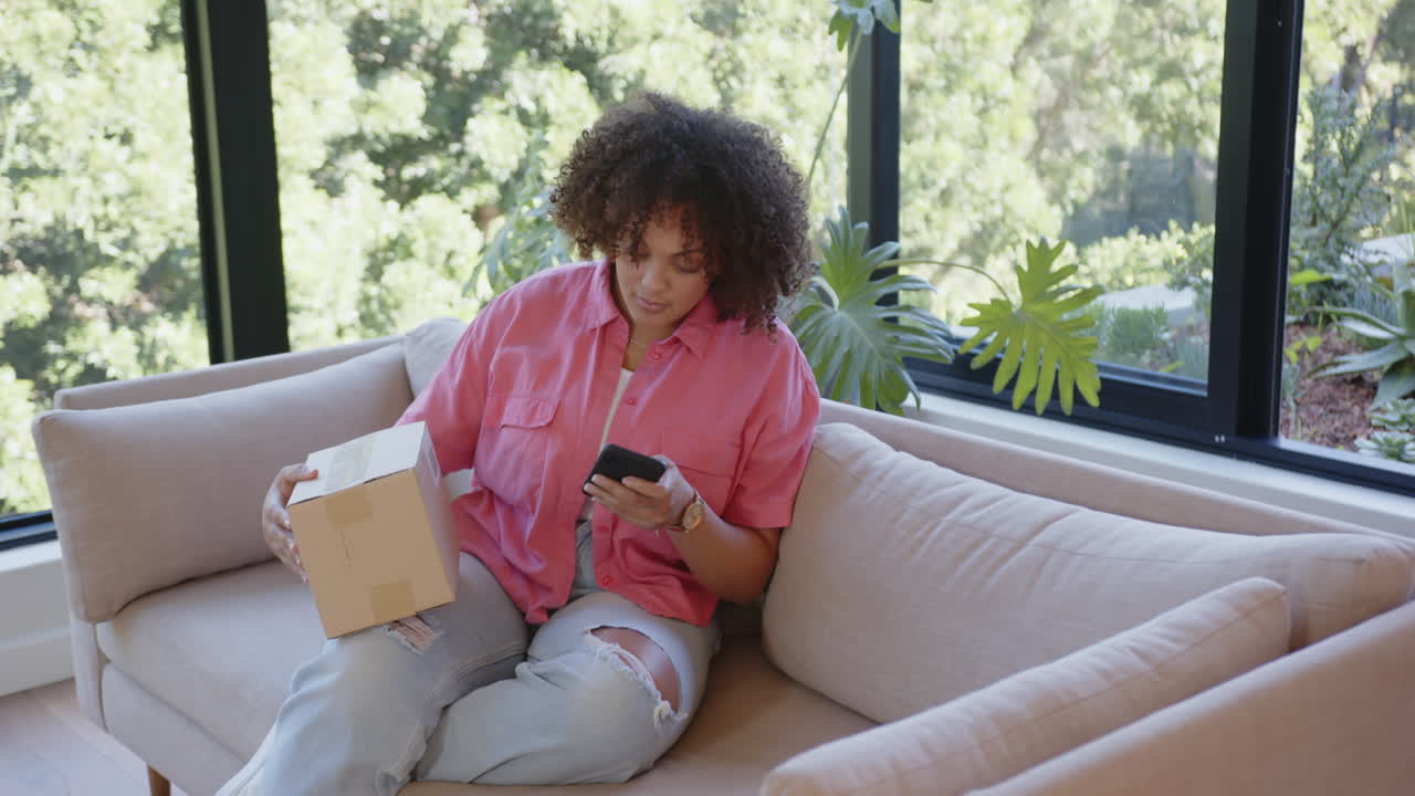 Sitting on couch, woman holding smartphone and package, checking delivery details