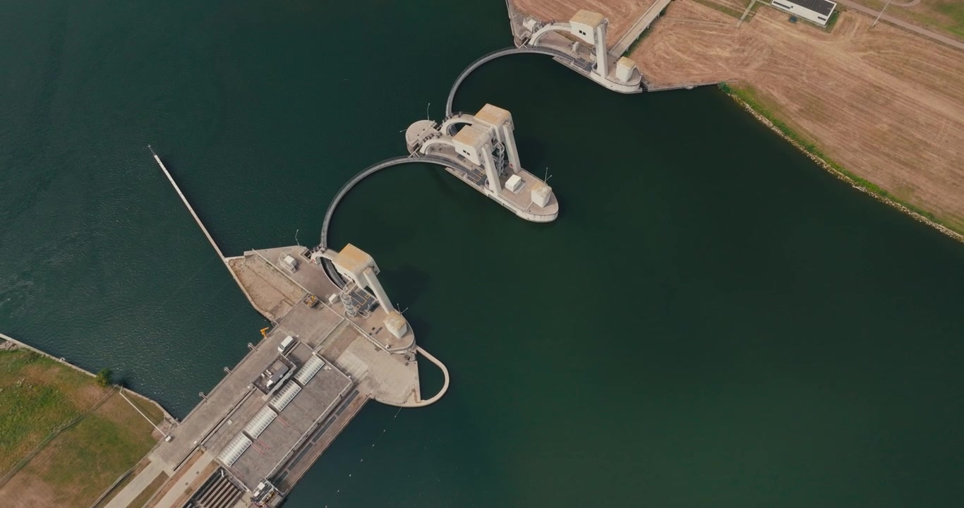 Drone flies away from the Maurik hydroelectric power plant on the Dutch Lower Rhine, revealing the complete engineering structure and distinctive semi-circular dam from above.