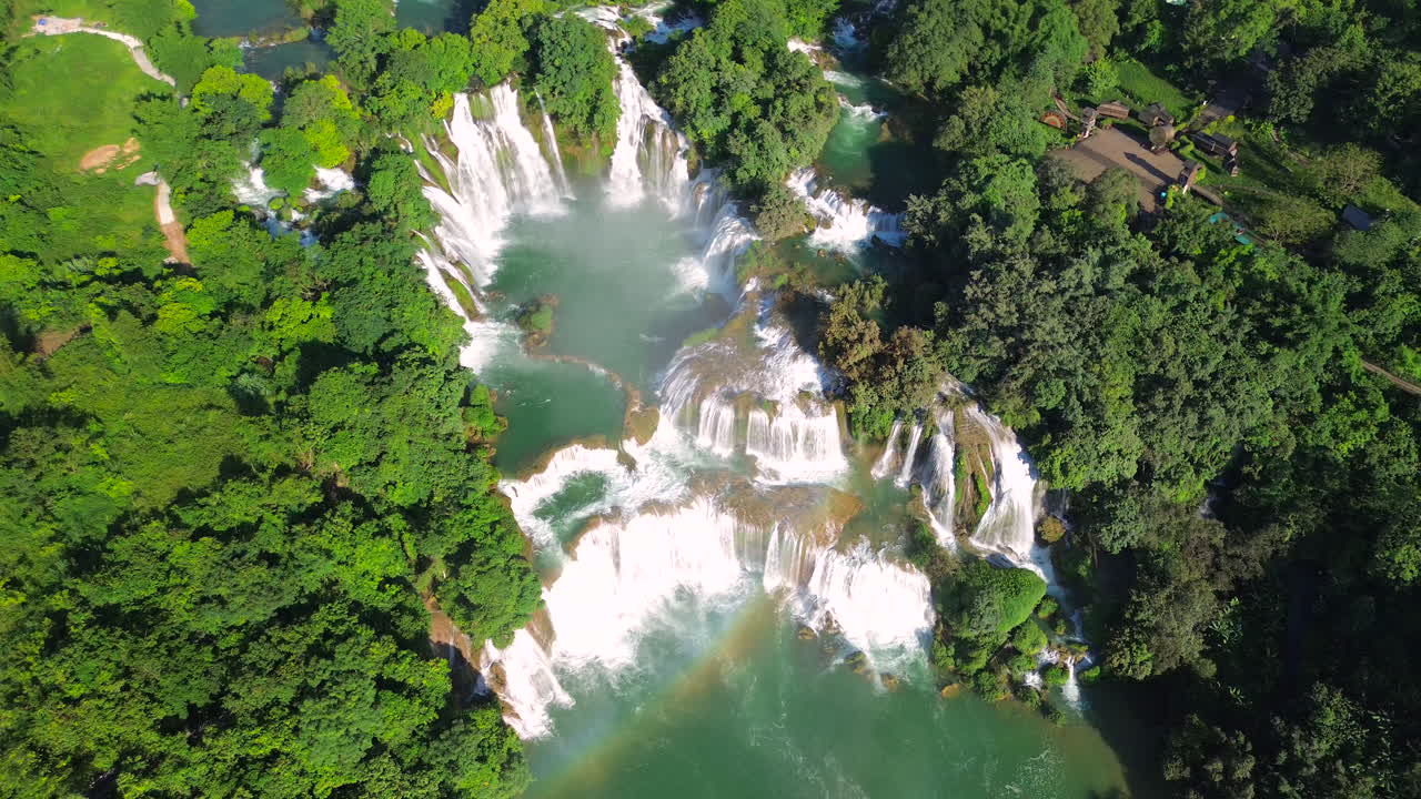 Scatto mozzafiato da drone della cascata a più livelli di Ban Gioc, con un arcobaleno visibile sull'acqua turchese e piccole imbarcazioni che navigano il fiume.
