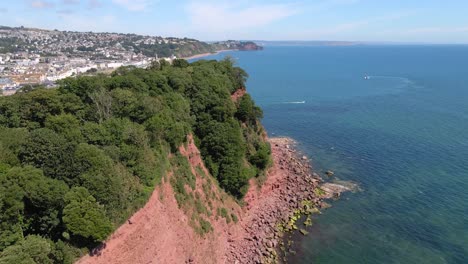 aerial flight along red cliff with trees on top and beautiful shaldon city in background