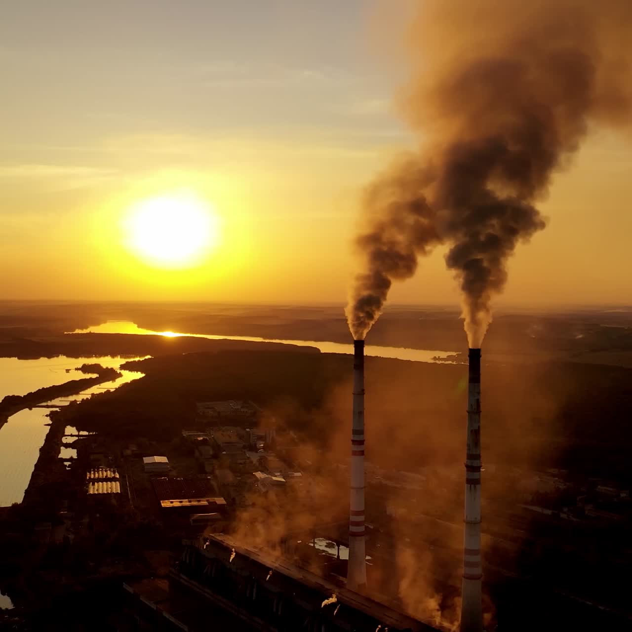 Metallurgy plant in nature at orange sunset. Dark smoke of industrial factory pollute the atmosphere. Evening background of a factory near the river.