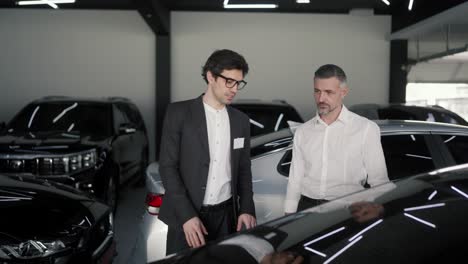 Un-Hombre-De-Mediana-Edad-Confiado,-Con-Barba-Gris-Y-Camisa-Blanca,-Se-Comunica-Con-Un-Joven-Moreno,-Un-Asistente-Con-Traje-De-Negocios-En-Un-Concesionario-De-Automóviles-Y-Examina-Un-Automóvil-Negro.