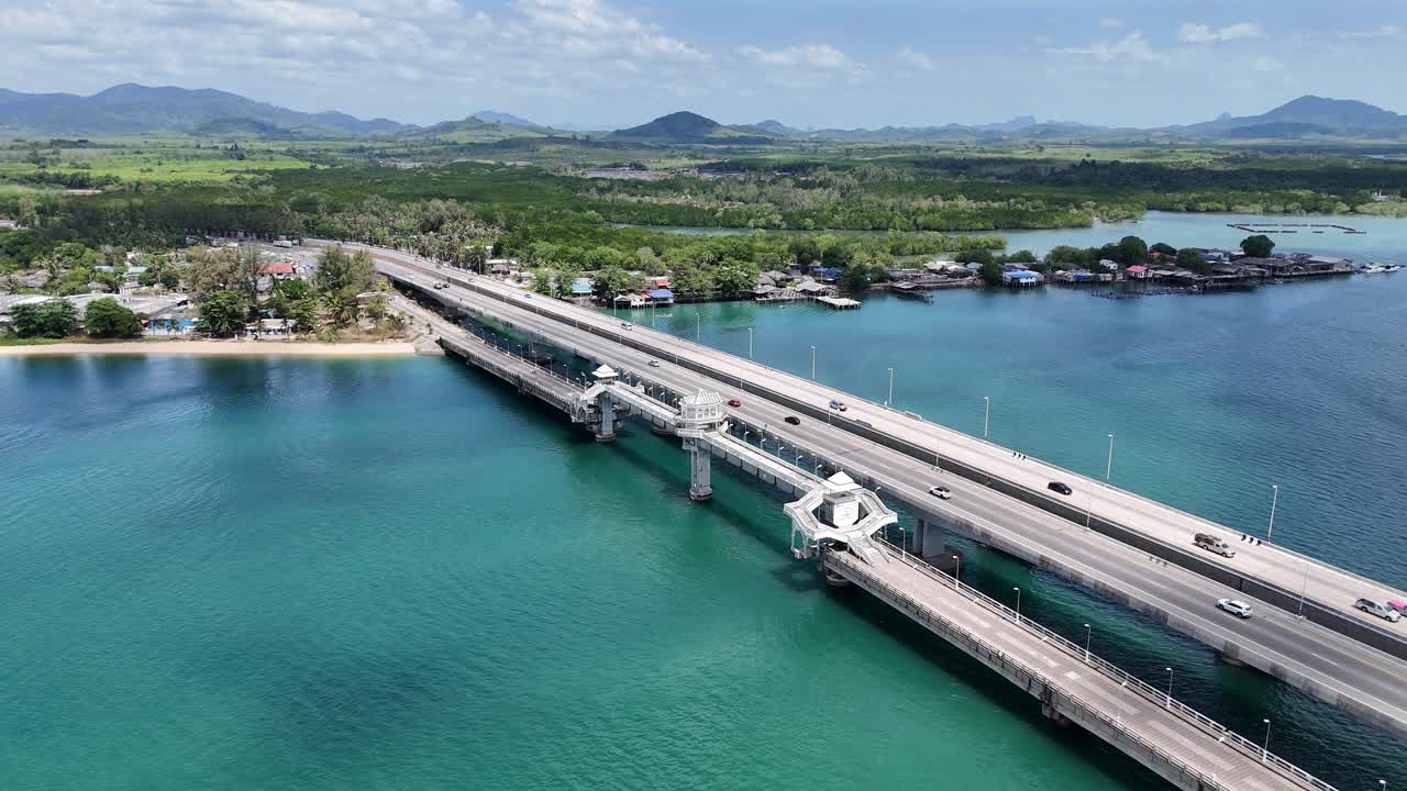 Drone footage captures Sarasin Bridge in Phuket, Thailand, spanning turquoise waters under clear skies. Vehicles traverse the scenic route