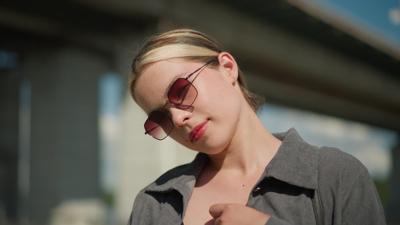 회색 셔츠를 입은 여자가 포즈를 취하는 동안 목걸이를 조정합니다. 그녀는 머리를 약간 기울여 자신감과 스타일을 발휘합니다. 흐릿한 배경에는 전기 기둥을 포함한 다리가 있습니다.