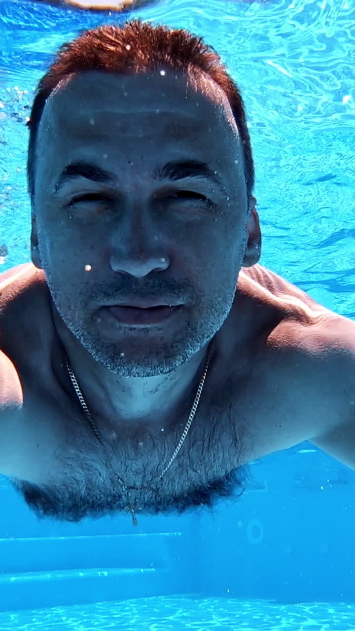 Man swimming underwater. Healthy man diving in the swimming pool and looking at camera. Happy male showing ok sign inside the pool. Vertical video