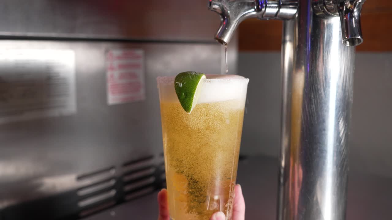 el camarero llena un vaso de pinta vestido con una rodaja de lima con cerveza de barril espumosa de un barril de acero inoxidable, cámara lenta 4k