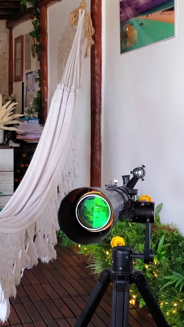 Telescope with a prominent green lens is set up outdoors, near white hammock and green plants. RElax and Education concept