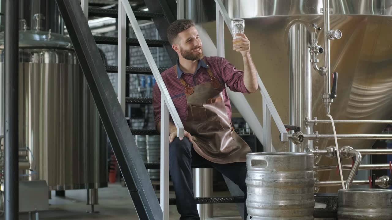 Young brewer wearing a leather apron is tasting beer at a modern brewery