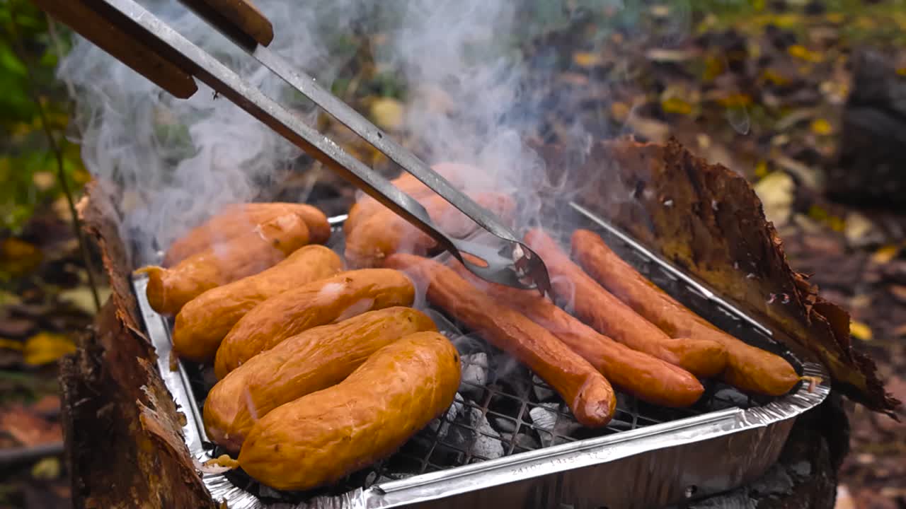 Golden brown and tasty sausages being cooked on a smoking hot disposable or instant grill BBQ in autumn nature with fallen yellow leaves and dirt in bokeh blurry background. Smoke move in slow motion