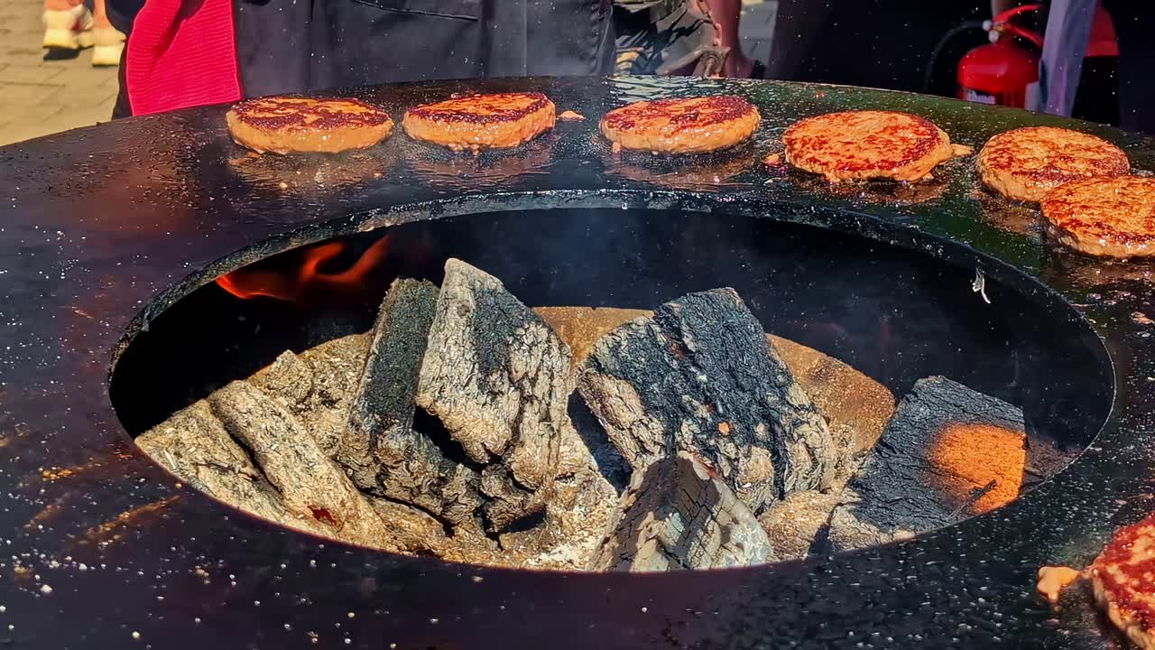 A close-up shot captures juicy beef burger patties sizzling on a large, round fire pit grill, with smoke and heat rising from the hot embers at a Latvian street food market