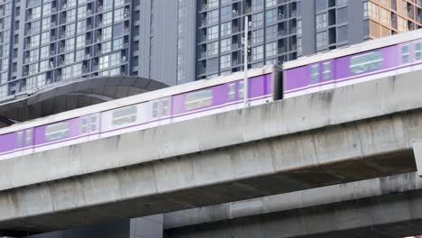 Lila-Zug,-Der-Von-Rechts-Nach-Links-Fährt-Und-An-Einem-Bahnhof-Mit-Hochhäusern-Im-Hintergrund-Hält,-Bangkok,-Thailand