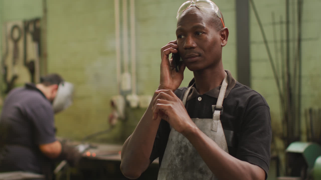 African American male factory worker at a factory standing in a workshop, talking on a smartphone