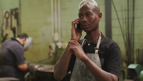 african american male factory worker at a factory standing in a workshop, talking on a smartphone