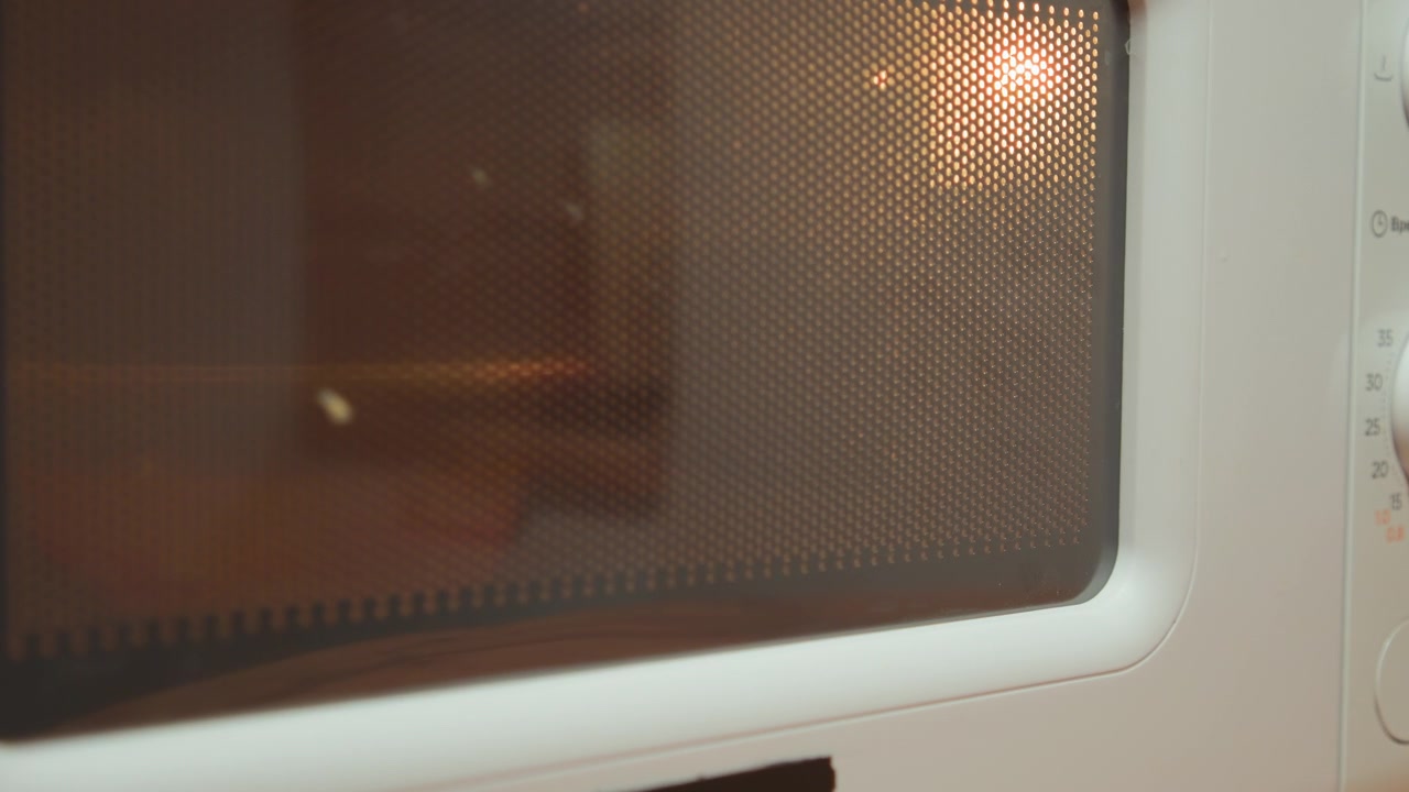 Primo piano frontale della porta di un forno a microonde con rete metallica perforata e vetro colorato che rivela un caldo bagliore interno ambrato, cibo in leggera rotazione, condensa sparsa e il riscaldamento quotidiano in cucina.