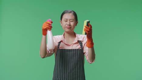 ama de casa asiática con un delantal mirando un spray y una esponja de cocina luego teniendo dolor de cabeza en el estudio de pantalla verde