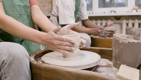 manos de empleados que usan delantal verde modelando piezas de cerámica en una rueda de alfarero en un taller