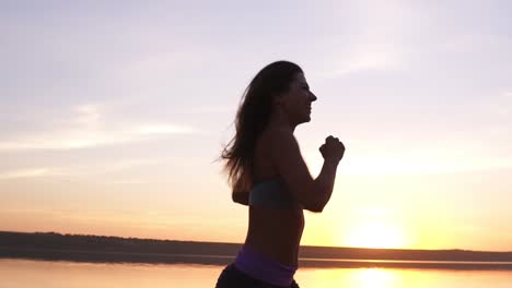 Close-up-footage-of-a-fit-girl-running-along-the-water.-Sunrise.The-young-woman-trainings.-Happy,-smiling