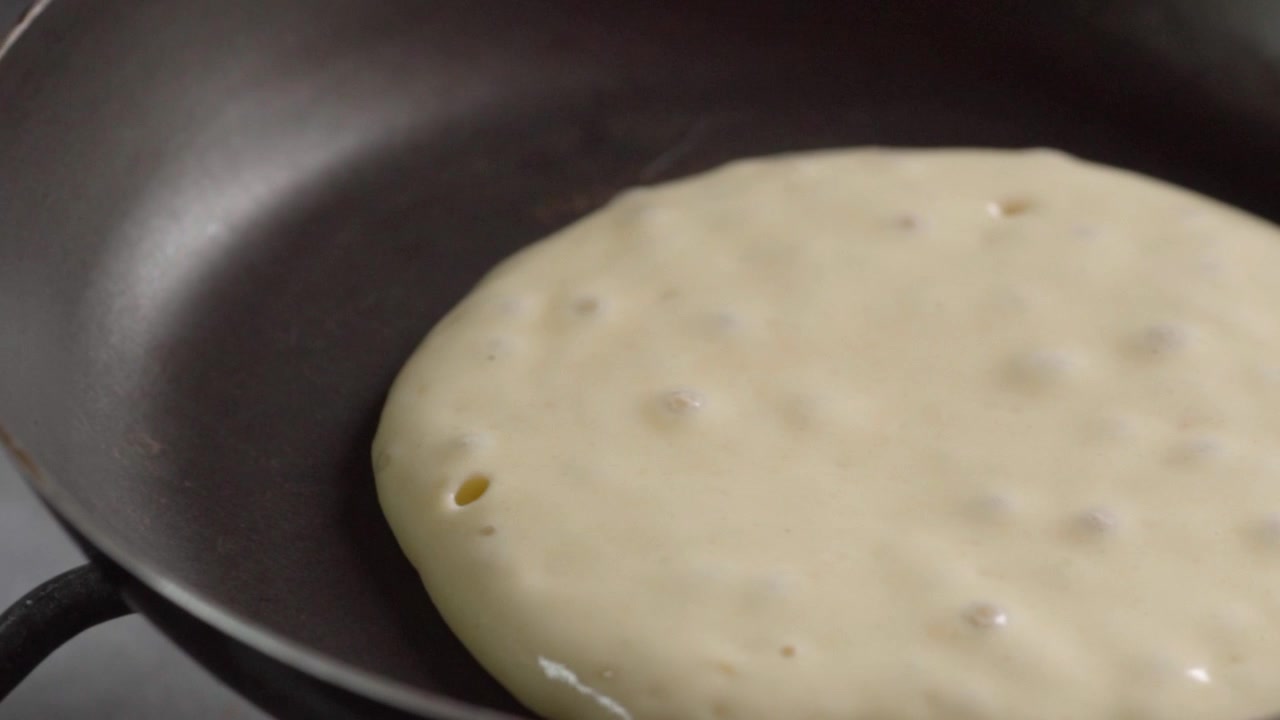 Close-up mostra massa de panqueca cozinhando em frigideira preta, com pequenas bolhas se formando em sua superfície enquanto começa a firmar durante o preparo do café da manhã.