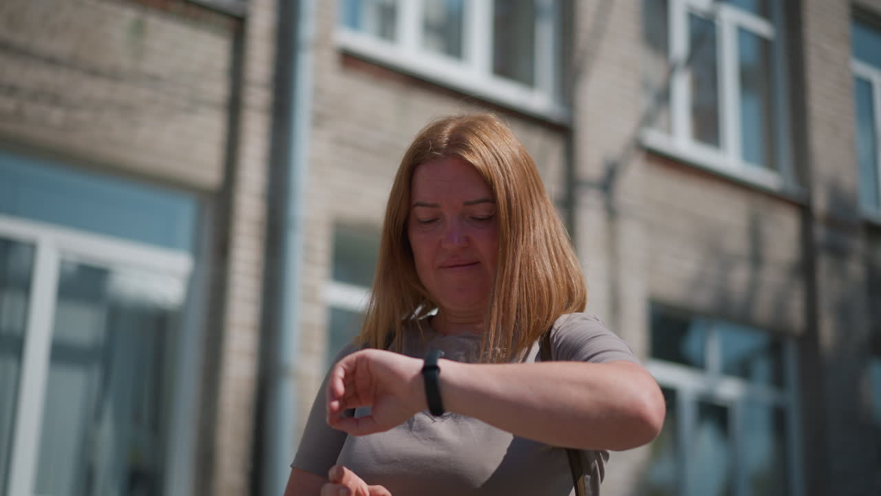 Sad woman standing under bright summer sunlight near building, looking around for someone and checking time with tired expression, sunlight reflecting on face, shadow visible on ground