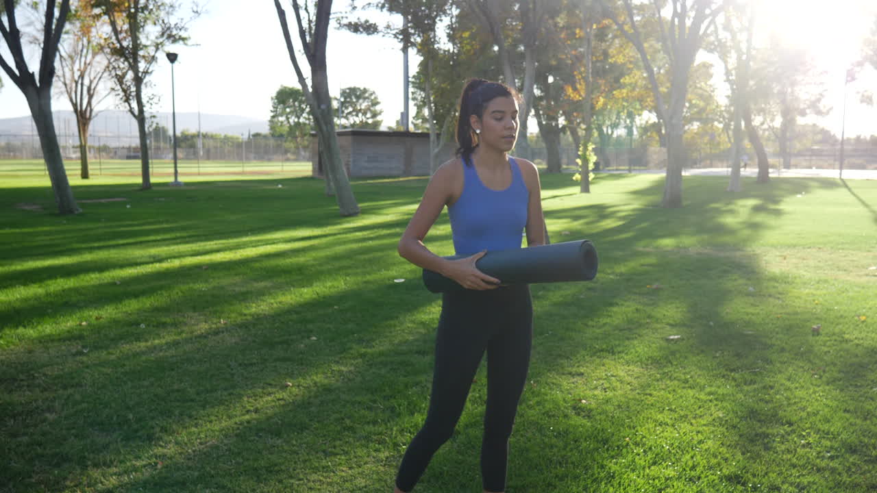 An attractive young hispanic woman rolling up her yoga fitness mat after a workout in the park at sunset SLOW MOTION
