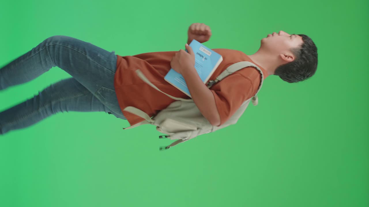 la vista lateral de un estudiante asiático con una mochila, una camiseta marrón y pantalones vaqueros azules sosteniendo el libro mientras corre a la escuela en la pantalla verde de chroma key.