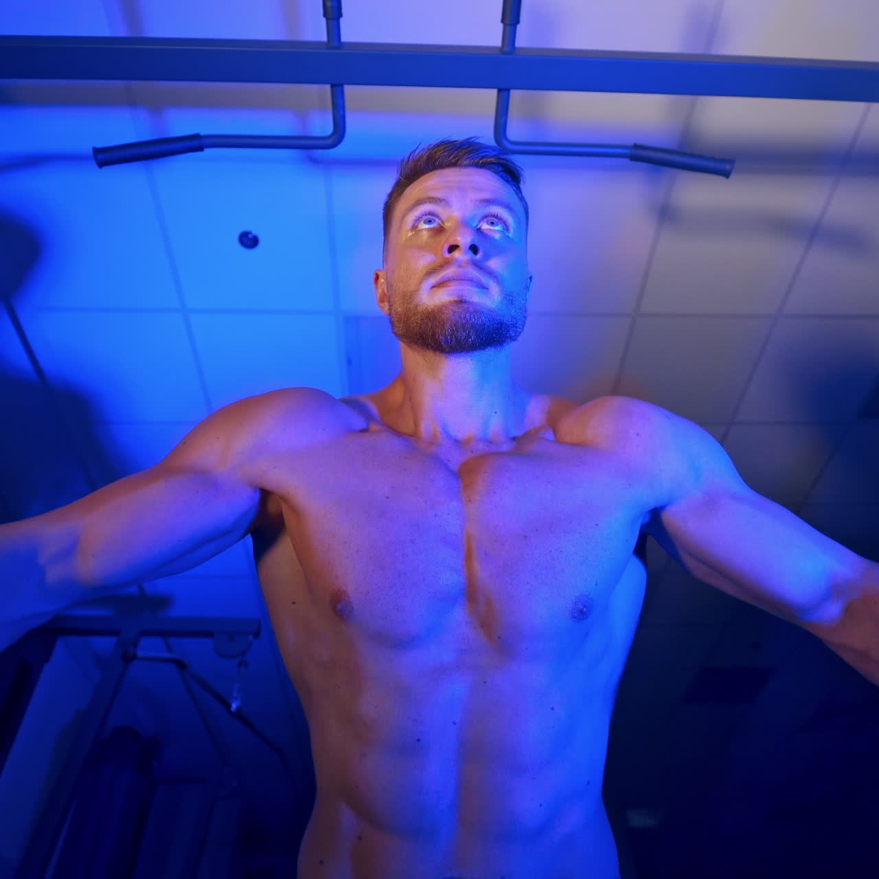 Handsome athlete pumps up his muscles. Naked torso of a sportsman. Muscular bodybuilder training in the gym. View from below. Blue light.