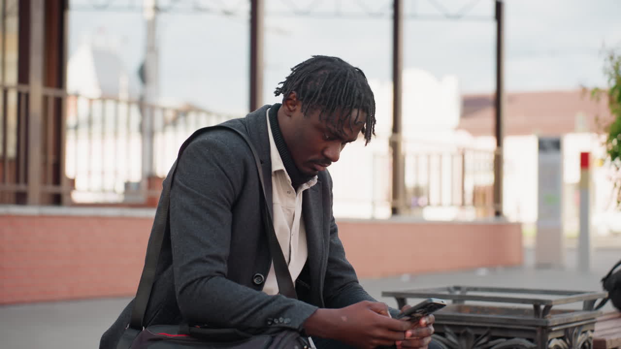 Dreadlocked man with short beard wearing layered clothing sits on wooden bench using smartphone outdoors, showing calm focus and stylish appearance in modern city with soft daylight reflections
